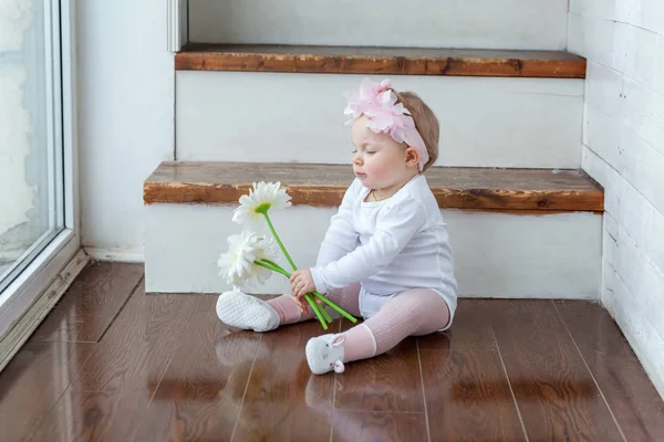 Petite fille portant une couronne de printemps assise sur le sol dans un salon lumineux près de la fenêtre et jouant avec des fleurs de gerbera — Photo