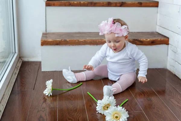 Niña pequeña con guirnalda de primavera sentada en el suelo en la sala de estar de luz brillante cerca de la ventana y jugando con flores de gerberas —  Fotos de Stock