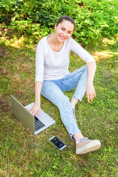 Молодая женщина, сидящая на лужайке в городском парке и работающая на ноутбуке. Концепция внештатного бизнеса — стоковое фото