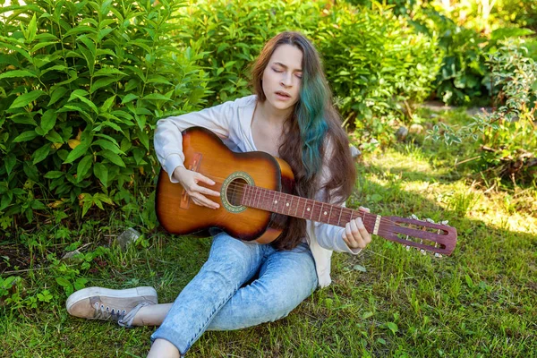 Młoda kobieta siedzi w trawie i gra na gitarze — Zdjęcie stockowe