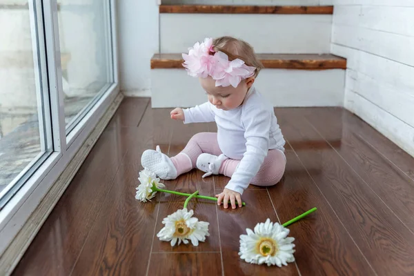 Lilla bebis flicka bär våren krans placering på golv i ljusa lätta vardagsrummet nära fönster och spelar med gerbera blommor — Stockfoto
