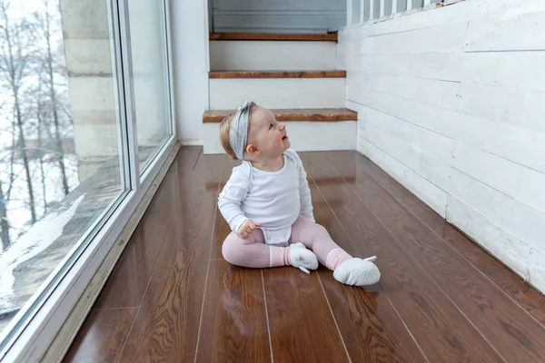 Little crawling baby girl one year old siting on floor in bright light living room near window smiling and laughing — Stock Photo, Image