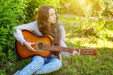 Genç hipster kadın çim oturan ve park veya bahçe arka plan üzerinde gitar çalıyor. Genç kız şarkı çalmayı öğrenme ve müzik yazma