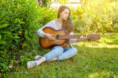 Genç hipster kadın çim oturan ve park veya bahçe arka plan üzerinde gitar çalıyor. Genç kız şarkı çalmayı öğrenme ve müzik yazma