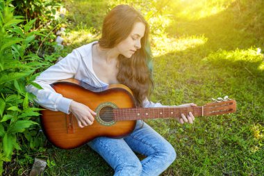 Genç hipster kadın çim oturan ve park veya bahçe arka plan üzerinde gitar çalıyor. Genç kız şarkı çalmayı öğrenme ve müzik yazma