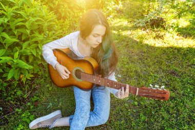Genç hipster kadın çim oturan ve park veya bahçe arka plan üzerinde gitar çalıyor. Genç kız şarkı çalmayı öğrenme ve müzik yazma