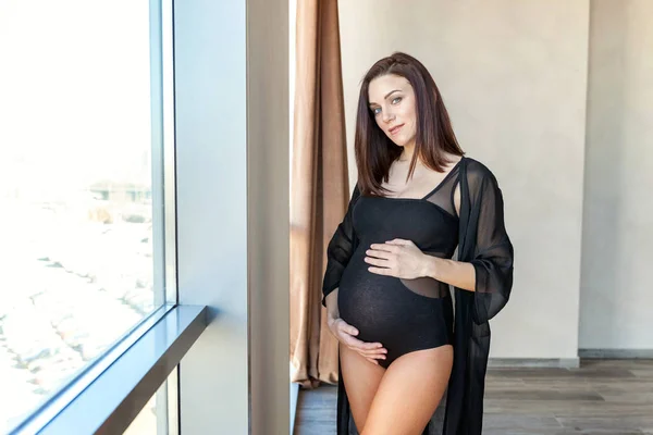 Těhotenství žena stojící poblíž okna doma a úsměvy — Stock fotografie