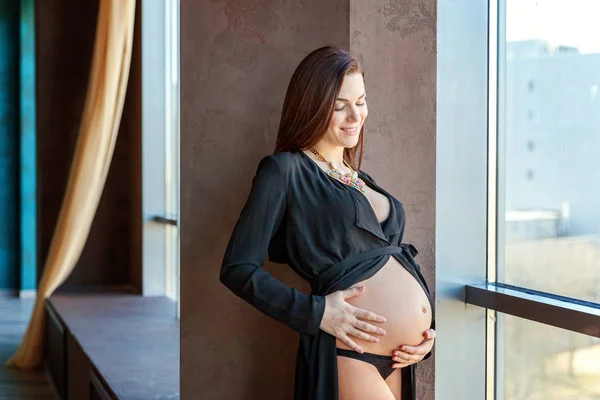 Mulher grávida de pé perto da janela em casa e sorri — Fotografia de Stock