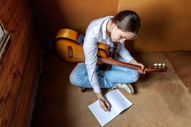 Young hipster woman sitting on floor and playing guitar at home clipart