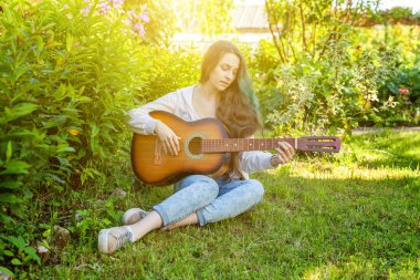 Genç hipster kadın çim oturan ve park veya bahçe arka plan üzerinde gitar çalıyor. Genç kız şarkı çalmayı öğrenme ve müzik yazma