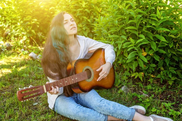 Young hipster woman sitting in grass and playing guitar on park or garden background. Teen girl learning to play song and writing music — Stock Photo, Image