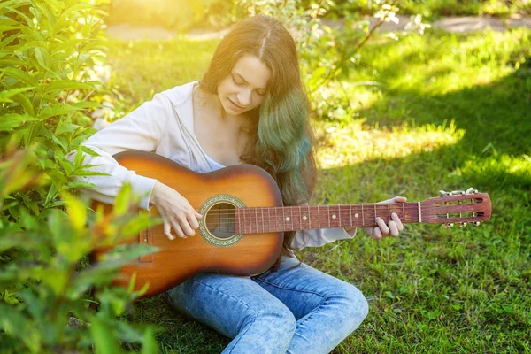 Young hipster woman sitting in grass and playing guitar on park or garden background. Teen girl learning to play song and writing music — Stock Photo, Image