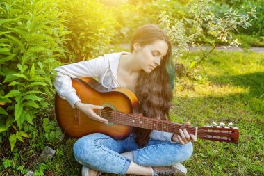 Genç hipster kadın çim oturan ve park veya bahçe arka plan üzerinde gitar çalıyor. Genç kız şarkı çalmayı öğrenme ve müzik yazma