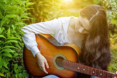 Young hipster woman sitting in grass and playing guitar on park or garden background. Teen girl learning to play song and writing music clipart
