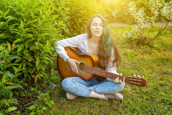 Joven mujer hipster sentado en la hierba y tocando la guitarra en el parque o jardín de fondo. Adolescente chica aprendiendo a tocar la canción y escribir música —  Fotos de Stock