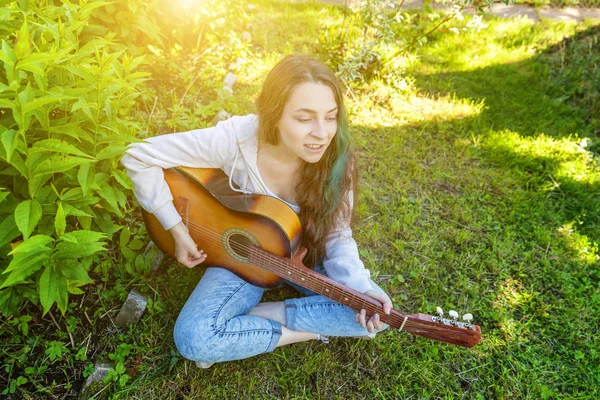 Fiatal csípő nő ül a fűben, és gitározni a parkban vagy a kert háttérben. Tini lány tanulás játszani dal és írás zene — Stock Fotó