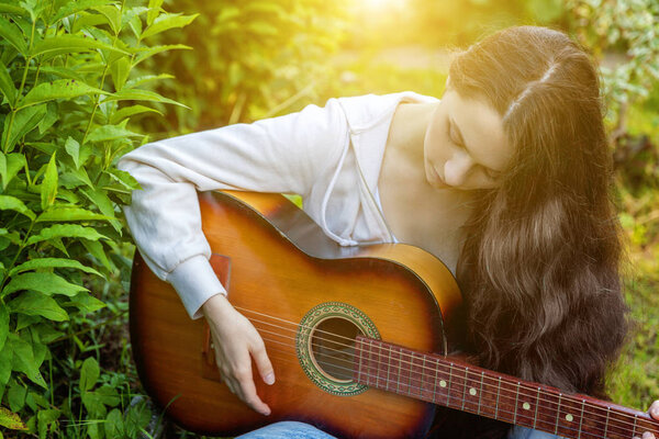 Young hipster woman sitting in grass and playing guitar on park or garden background. Teen girl learning to play song and writing music