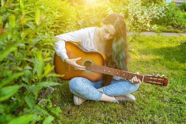 Jonge hipster vrouw zittend in gras en het spelen van gitaar op Park of tuin achtergrond. Tiener meisje leert liedjes te spelen en muziek te schrijven — Stockfoto