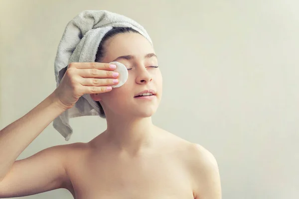 Smiling woman in towel on head with soft healthy skin removing make up with cotton pad isolated on white background