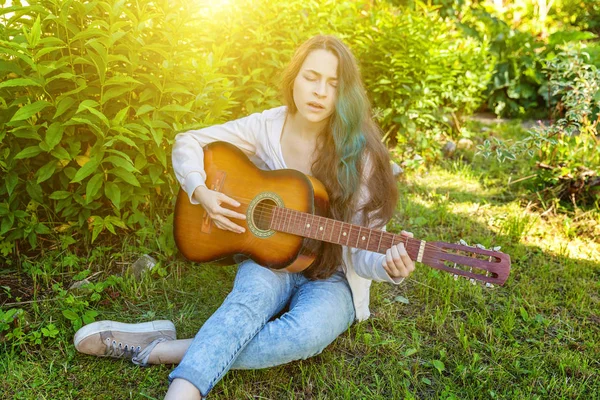 Mulher jovem hipster sentado na grama e tocando guitarra no parque ou jardim de fundo. Adolescente aprendendo a tocar música e escrever música — Fotografia de Stock