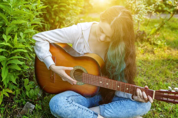 Joven mujer hipster sentado en la hierba y tocando la guitarra en el parque o jardín de fondo. Adolescente chica aprendiendo a tocar la canción y escribir música —  Fotos de Stock