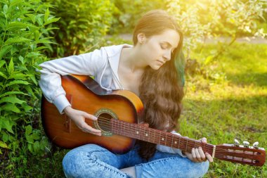 Genç hipster kadın çim oturan ve park veya bahçe arka plan üzerinde gitar çalıyor. Genç kız şarkı çalmayı öğrenme ve müzik yazma