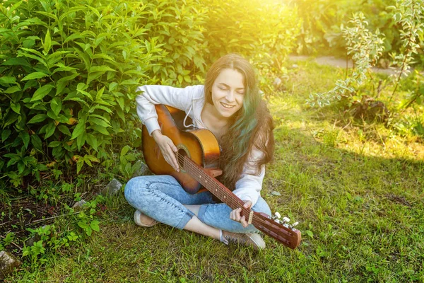 Mladá hipíků sedí v trávě a hraje na kytaru v parku nebo v zahradě. Dívka dospívajících pro hraní písní a psaní hudby — Stock fotografie