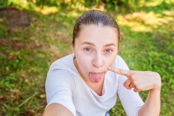 Mladá dívka si bere Selži z rukou s telefonem zobrazující jazyk a legrační obličej, který sedí na parku nebo na zahradě — Stock fotografie