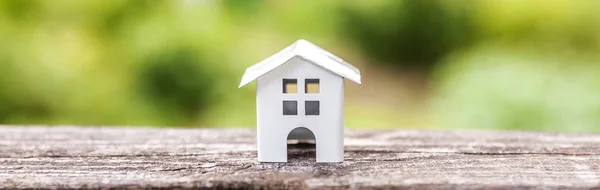 Miniature white toy model house in wooden background near green backdrop Banner