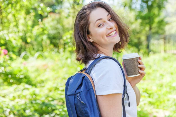 Gelukkig positief student meisje met rugzak en take-away koffie kopje glimlachend op groen park achtergrond — Stockfoto