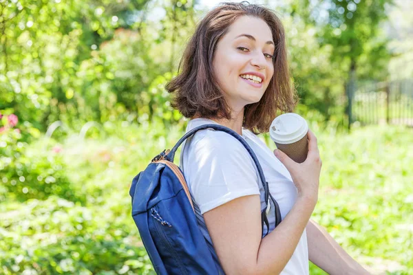 Heureuse étudiante positive avec sac à dos et emporter tasse de café souriant sur fond de parc vert — Photo