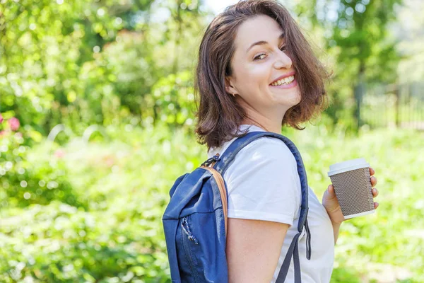 Sırt çantası ile mutlu pozitif öğrenci kız ve yeşil Park arka planda gülümseyen kahve bardağı almak
