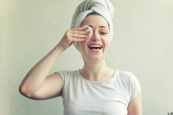 Mulher sorridente em toalha na cabeça com pele macia e saudável removendo maquiagem com almofada de algodão isolada no fundo branco — Fotografia de Stock