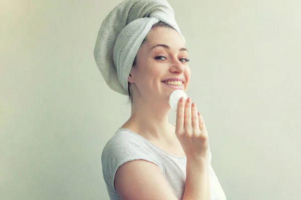 Mulher sorridente em toalha na cabeça com pele macia e saudável removendo maquiagem com almofada de algodão isolada no fundo branco — Fotografia de Stock