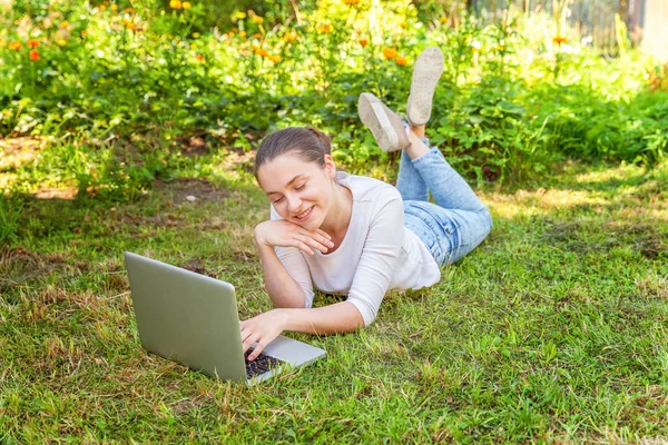Молодая женщина, лежащая на лужайке в городском парке и работающая на ноутбуке. Концепция внештатного бизнеса — стоковое фото