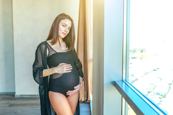 Těhotenství žena stojící poblíž okna doma a úsměvy — Stock fotografie