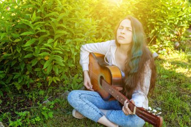 Genç hipster kadın çim oturan ve park veya bahçe arka plan üzerinde gitar çalıyor. Genç kız şarkı çalmayı öğrenme ve müzik yazma
