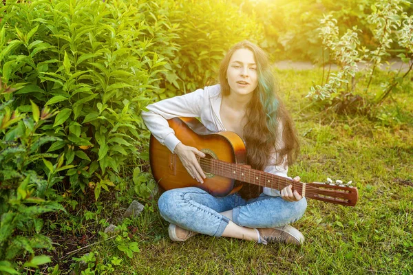 Mulher jovem hipster sentado na grama e tocando guitarra no parque ou jardim de fundo. Adolescente aprendendo a tocar música e escrever música — Fotografia de Stock