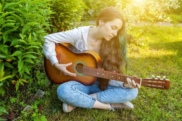 Jonge hipster vrouw zittend in gras en het spelen van gitaar op Park of tuin achtergrond. Tiener meisje leert liedjes te spelen en muziek te schrijven — Stockfoto