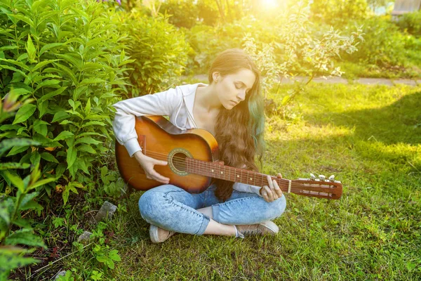 Молода хіпстерка сидить у траві і грає на гітарі на фоні парку або саду. Дівчина-підліток вчиться грати пісню і писати музику — стокове фото