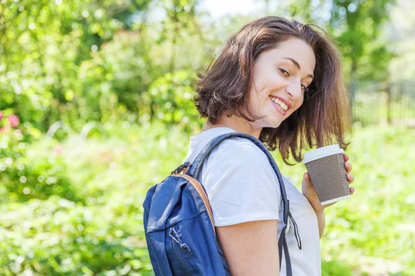 Sırt çantası ile mutlu pozitif öğrenci kız ve yeşil Park arka planda gülümseyen kahve bardağı almak