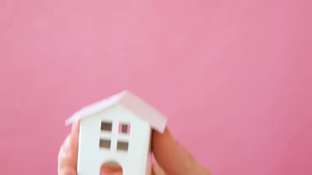 ピンクのパステルカラフルなトレンディな背景に隔離されたミニチュア白いおもちゃの家を保持する単に女性の女性の手を設計 — ストック動画