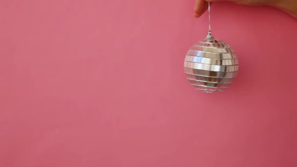 ピンクのパステルカラーのトレンディーな背景に隔離されただけで最小限のデザインの女性の手はクリスマスオーナメントディスコボールを保持 クリスマスの新年のお祝いの概念のための12月の時間 スペースのコピー — ストック動画
