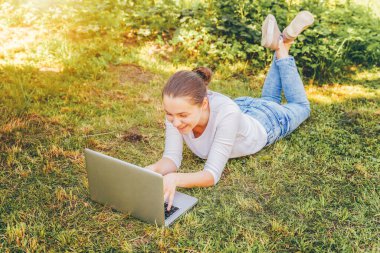Mobil Ofis. Serbest iş konsepti. Şehir parkında çimlerin üzerinde yatan genç bir kadın dizüstü bilgisayarla çalışıyor. Yaşam tarzı otantik, açık havada okuyan samimi öğrenci kız..