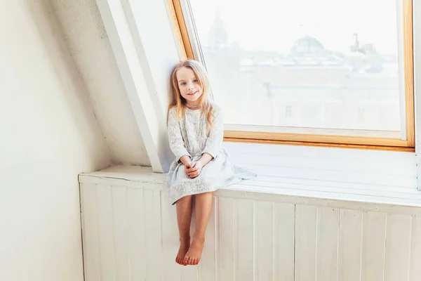 Little cute sweet smiling girl in white dress sitting on window sill in bright light living room at home indoors. Childhood schoolchildren youth relax concept