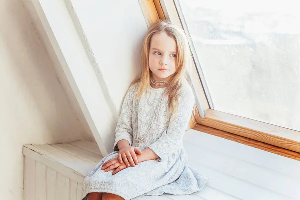 Little cute sweet smiling girl in white dress sitting on window sill in bright light living room at home indoors. Childhood schoolchildren youth relax concept