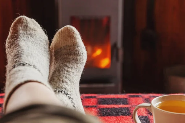 Feet legs in winter clothes wool socks and cup tea at fireplace background. Woman sitting at home on winter or autumn evening relaxing and warming up. Winter and cold weather concept. Hygge Christmas eve