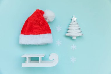 Kışlık nesnelerin çok az bileşimi Noel Baba 'nın Noel Baba şapkalı kızağı köknar ağacını süsler mavi pastel renkli arka planda izole eder. Noel, yeni yıl kutlamaları için aralık zamanı. Düz üst görünüm