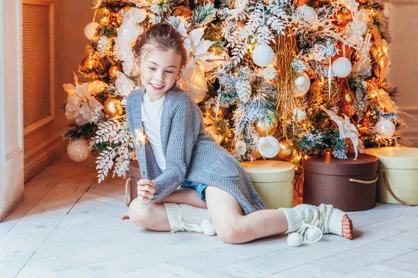 Κοριτσάκι με σπίθα κοντά στο χριστουγεννιάτικο δέντρο παραμονή Χριστουγέννων στο σπίτι. Νεαρό παιδί στο φως υπνοδωμάτιο με διακόσμηση χειμώνα Ευτυχισμένη οικογένεια στο σπίτι Χριστούγεννα Πρωτοχρονιά Δεκέμβριος χρόνο για εορτασμό έννοια — Φωτογραφία Αρχείου