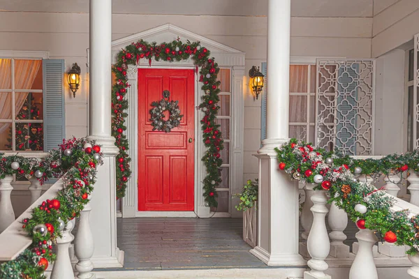 Idéia da decoração do alpendre do Natal. Entrada da casa com porta vermelha decorada para férias. grinalda grinalda vermelha e verde de ramos de abeto e luzes sobre trilhos. Véspera de Natal em casa — Fotografia de Stock